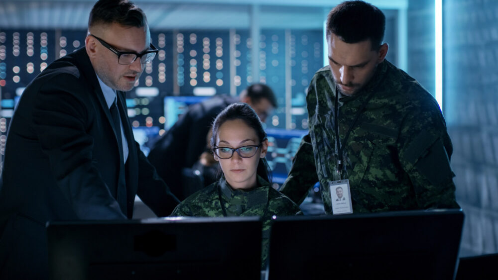 Three individuals, two in military uniforms and one in a suit, are working at computer screens in a government control room equipped with custom technology, against a backdrop of monitors and data servers.