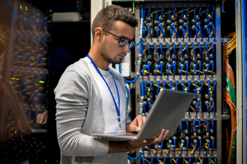 A man in a gray cardigan uses a laptop while standing in front of a server rack filled with numerous cables, showcasing the efficiency of Ace Computers CCS-2 2019B products in a data center.