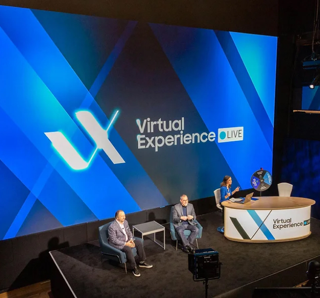 Three people sit on a stage with a large screen displaying "Samsung V/X Live" at a professional event.