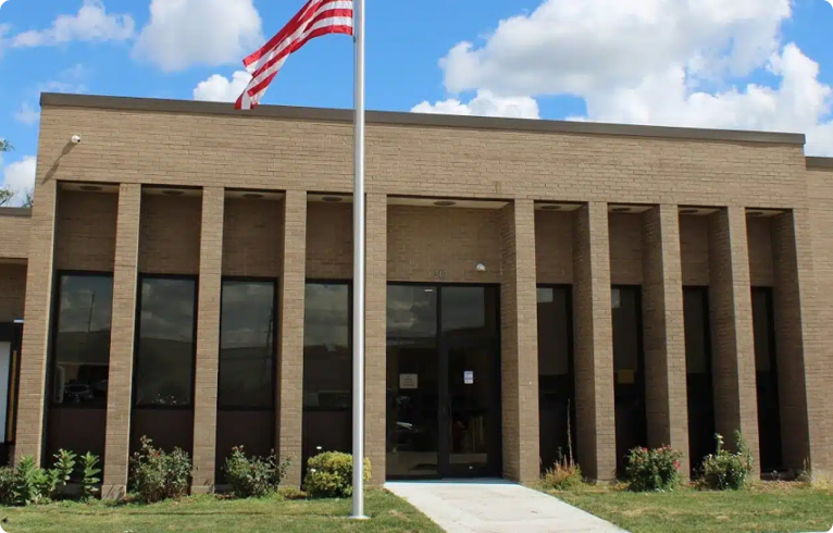 Brick building with dark windows under a blue sky, featuring tall pillars and an American flag flying on a pole at the front of the new home.