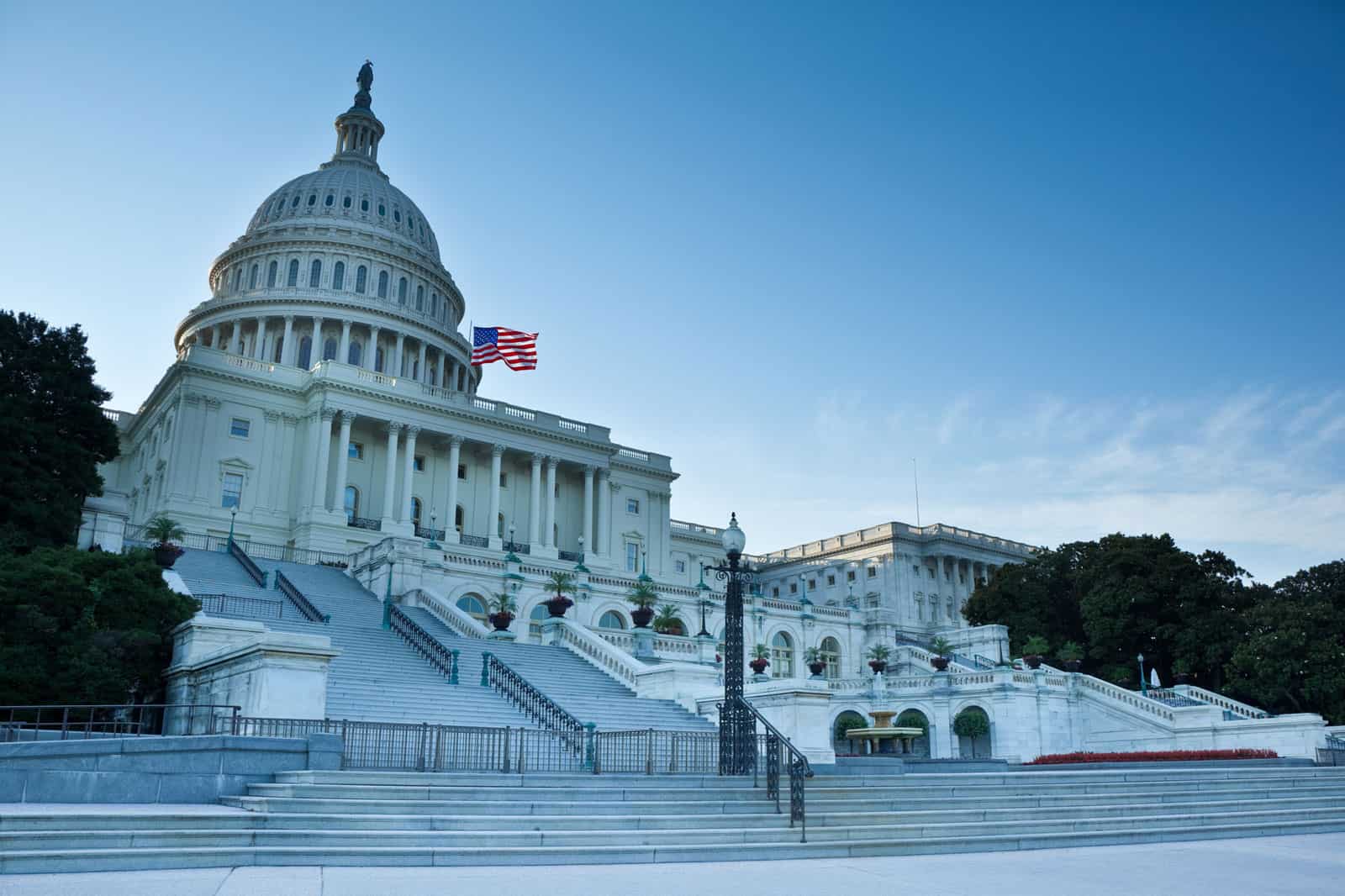 The United States Capitol building stands majestic, its iconic dome and an American flag soaring against a clear blue sky. Stone steps and lampposts—essential features of its architecture—grace the foreground.