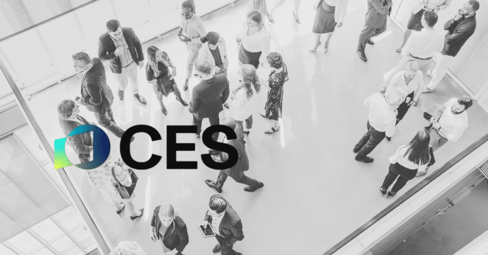 Overhead view of a group of people networking in a bright, modern indoor space with the CES and I/ITSEC 2025 logos prominently displayed in the foreground.