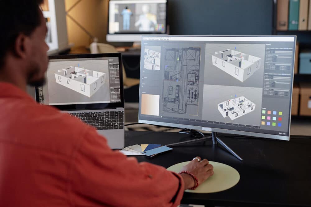 Person working at a desk with a laptop and monitor displaying 3D architectural floor plans and renderings in design software, utilizing Computational Fluid Dynamics for advanced analysis.