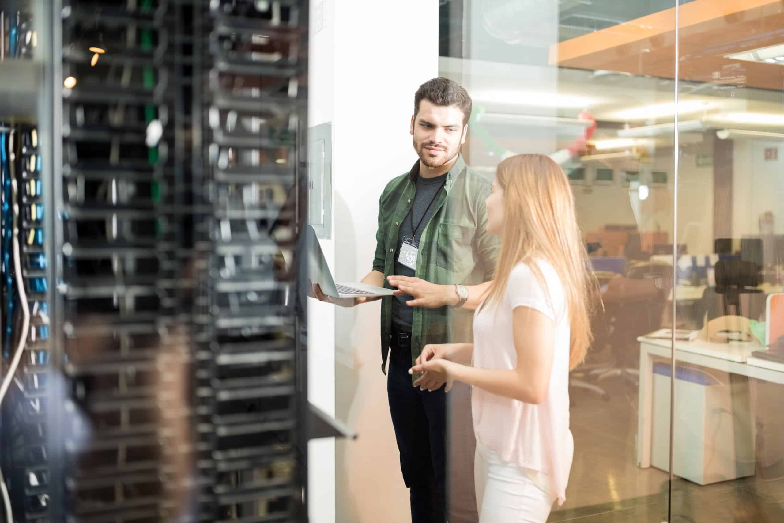 Two people stand near a glass wall in an office, discussing something about the Corvid HPC partnership while one holds a laptop next to server racks.