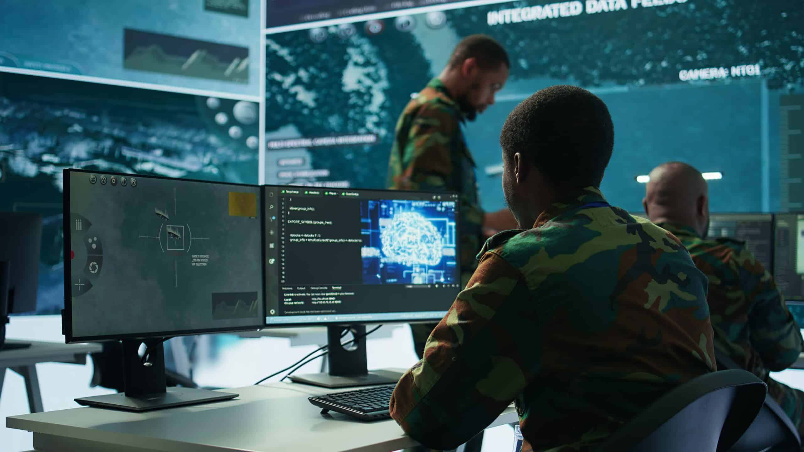 Military personnel in camouflage uniforms work at computer stations displaying surveillance data and maps, utilizing advanced Army Solutions in a high-tech operations room.