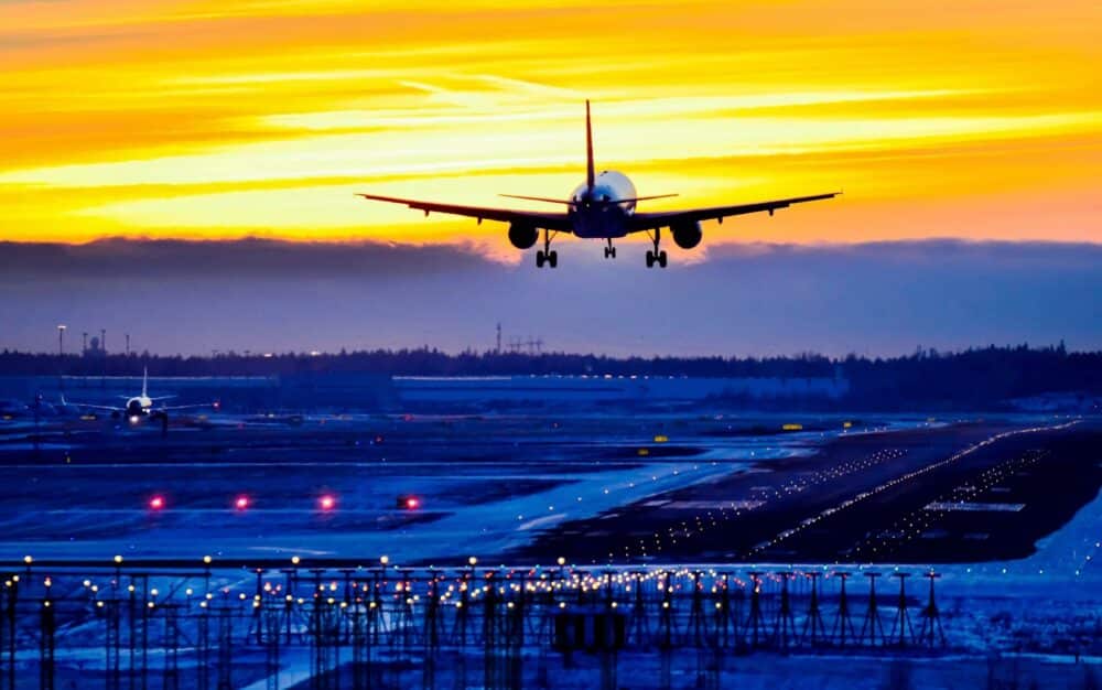 A commercial airplane approaches a runway for landing at sunset, with runway lights illuminated and another plane visible in the background.