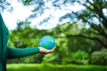Person holding a small globe model in their hand with a blurred green forest background, symbolizing global efforts like EPEAT Climate+ initiatives for environmental sustainability.