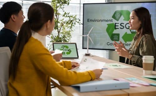 Three people sit at a table discussing ESG (Environmental, Social, Governance) strategies in a modern office, with a presentation screen highlighting the benefits of low-power GPUs and documents spread out on the table.