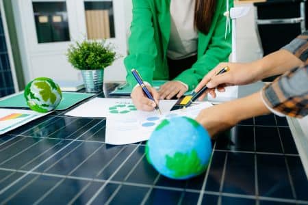 Two people work on documents with recycling symbols at a desk featuring solar panels and model globes, highlighting a focus on environmental sustainability and EPEAT Gold standards.