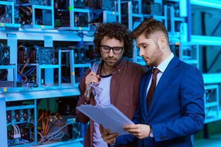 Two men stand in a server room or data center; one, in casual clothes with a coiled cable, and the other, suited and reviewing documents near ENERGY STAR certified equipment.