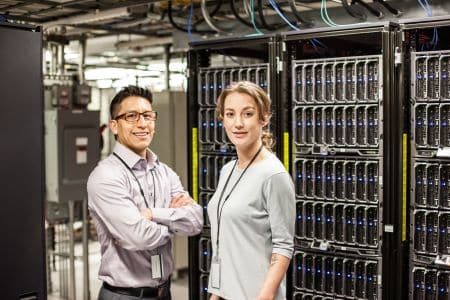 Two people stand in a server room filled with HPC servers and computer equipment. Both are wearing office attire and visible employee ID badges.
