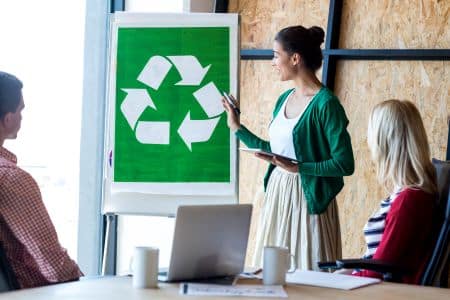A woman is presenting a large recycling symbol and highlighting EPEAT Gold benefits on a flip chart to two seated colleagues in a meeting room.