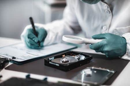 Person in lab attire examining an open hard drive with a magnifying glass, recording notes on a clipboard, and using a USB write blocker for secure forensic analysis.