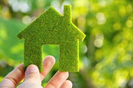 A hand holds a small house-shaped cutout covered in green grass-like material, symbolizing epeat climate+ and ENERGY STAR certification, with greenery and sunlight blurred in the background.