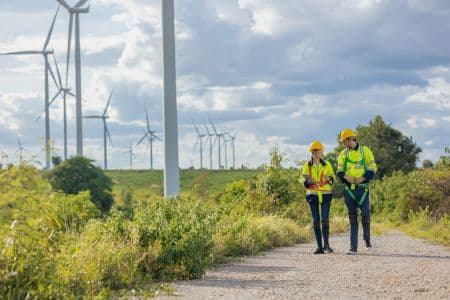 Two engineers in safety gear walk on a path near ENERGY STAR Certified wind turbines in a rural landscape under a cloudy sky.
