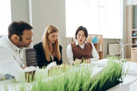 Three people sit at a table in an office, discussing documents. A model wind turbine and green plants highlight their focus on ESG initiatives and AI Sustainability in the foreground.
