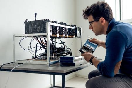 A man in glasses examines a graphics card beside a cryptocurrency mining rig with ENERGY STAR certification on a table in a minimally furnished room.