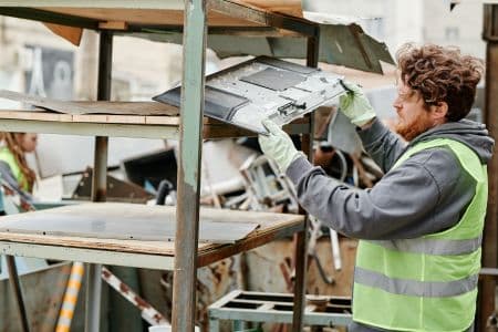 A man wearing a safety vest and gloves sorts metal scrap on shelves at a recycling facility, supporting epeat gold standards for sustainable materials management.