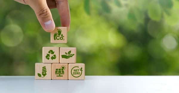 A hand stacks wooden blocks with eco-friendly icons, including CO2, recycling, renewable energy, plant, and net zero symbols—highlighting sustainable solutions like low-power GPUs—against a blurred green background.