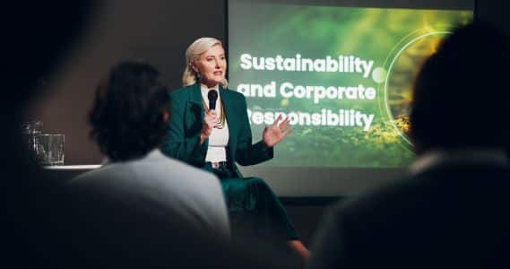 A woman in a green suit speaks into a microphone in front of a screen displaying the text "Sustainability and Corporate Responsibility" to an audience, highlighting the importance of ENERGY STAR Certified practices.