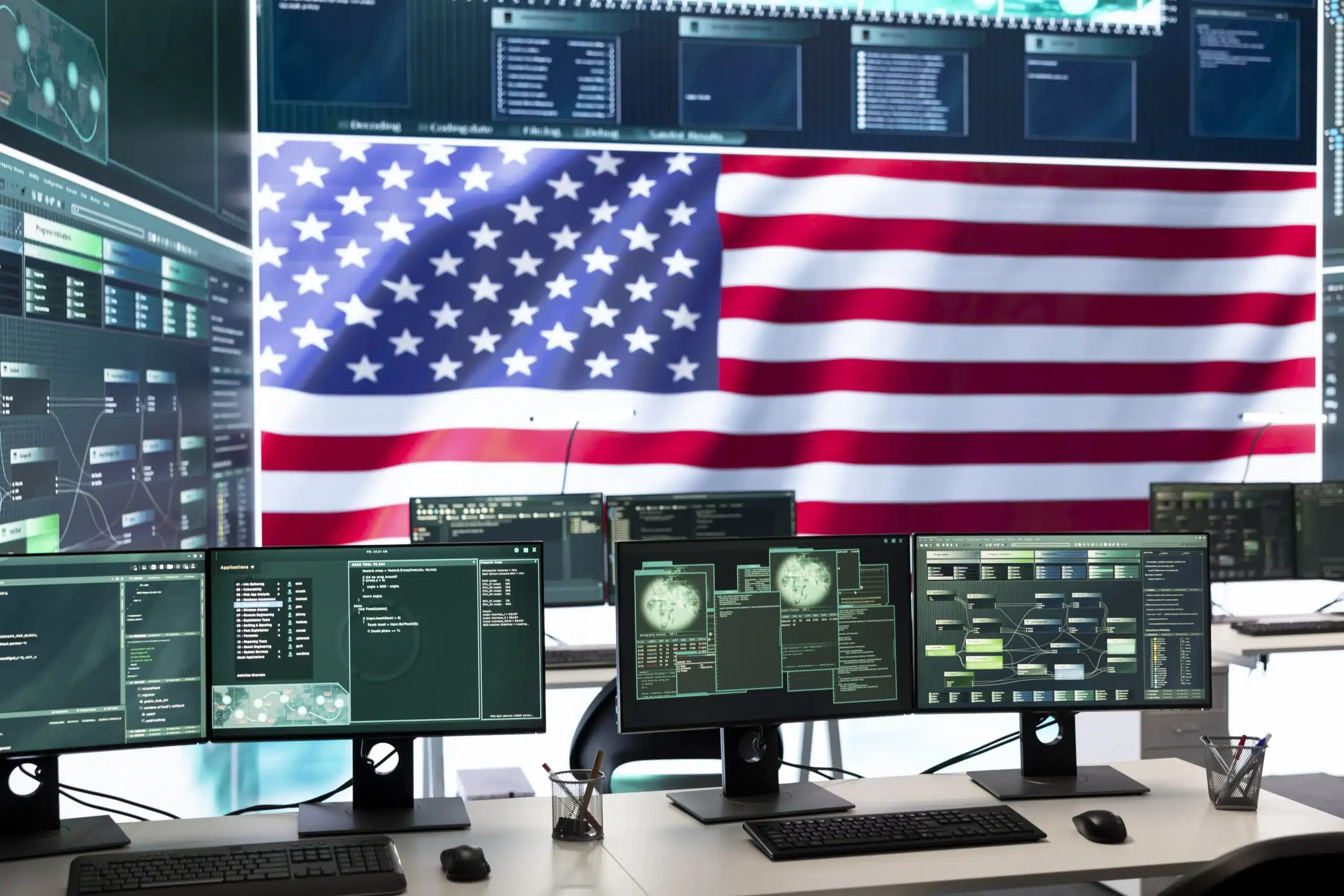 Computer workstations display federal and military data and analytics in a control room, with a large United States flag visible in the background.