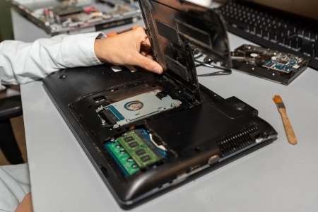 A person repairs a laptop with the back cover removed, exposing internal components and checking connections, possibly for RAID or NAS setup, on a gray desk.