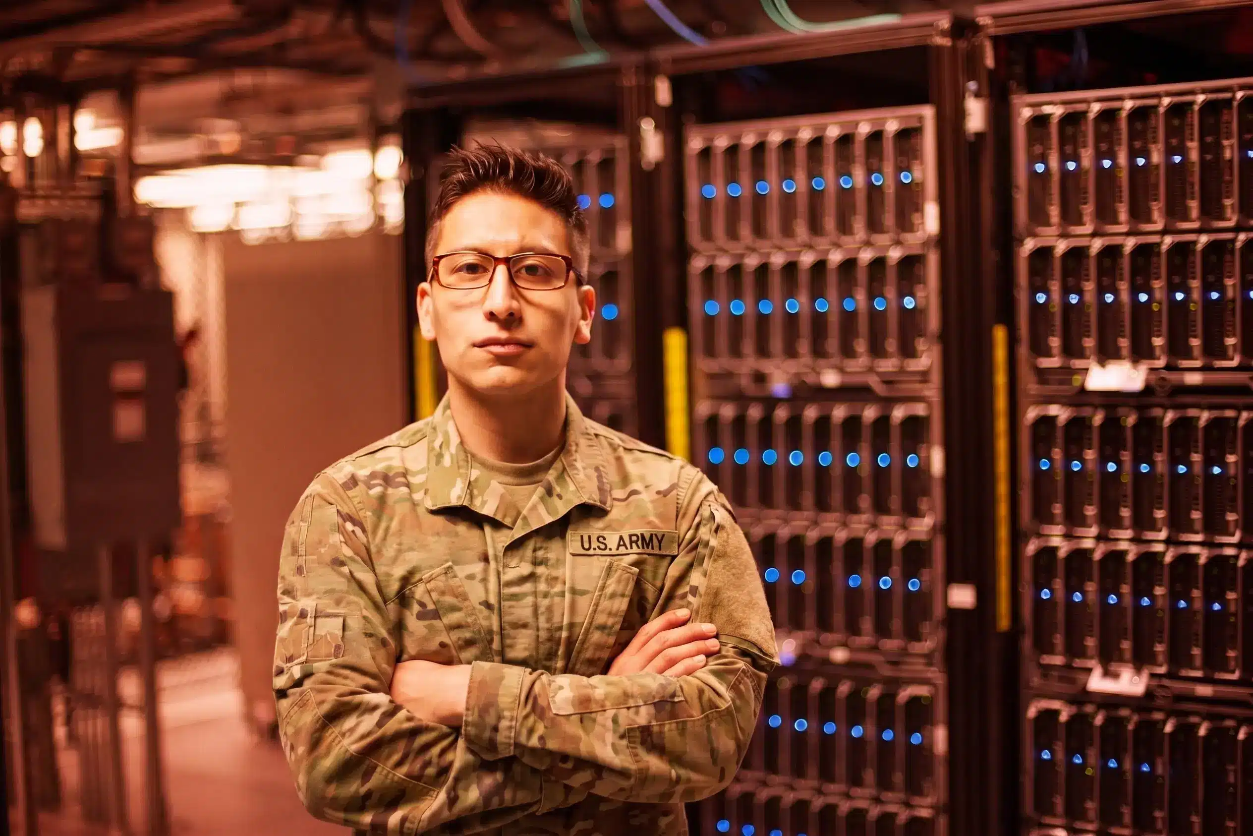 A person in a U.S. Army uniform stands with arms crossed in front of server racks in a federal or military data center.