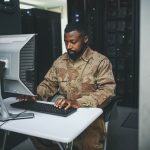 A man in military uniform works on a computer at a small desk surrounded by tall racks of servers, showcasing the cutting-edge infrastructure of computers for military applications.