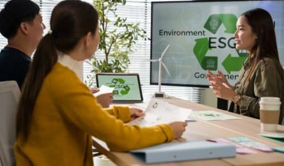 Three people sit at a table discussing ESG (Environmental, Social, Governance) strategies in a modern office, with a presentation screen highlighting the benefits of low-power GPUs and documents spread out on the table.