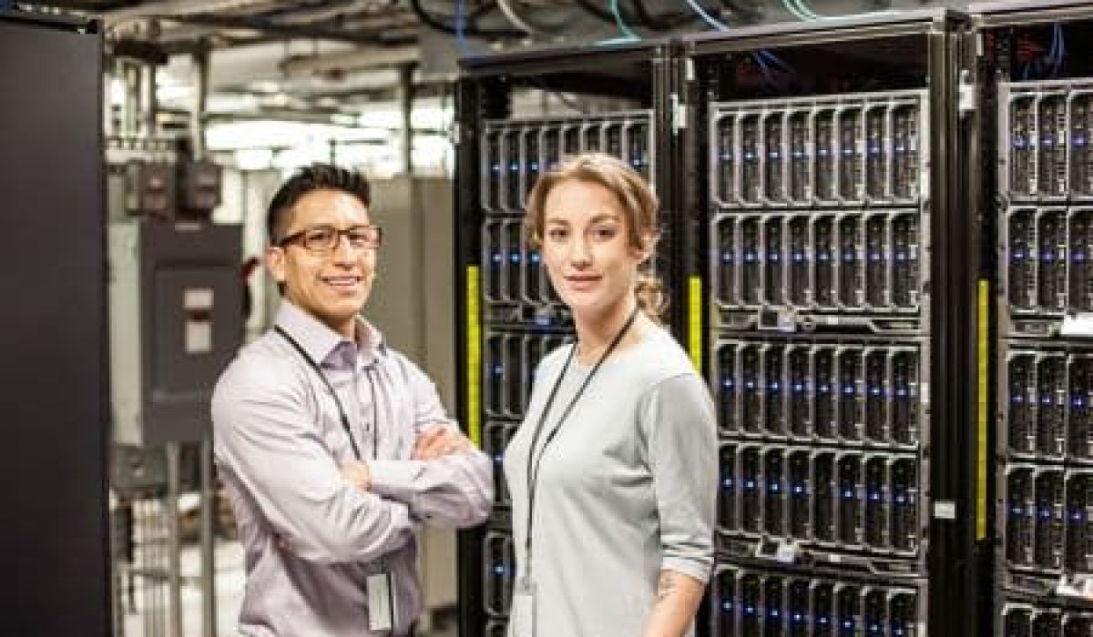 Two people stand in a server room filled with HPC servers and computer equipment. Both are wearing office attire and visible employee ID badges.