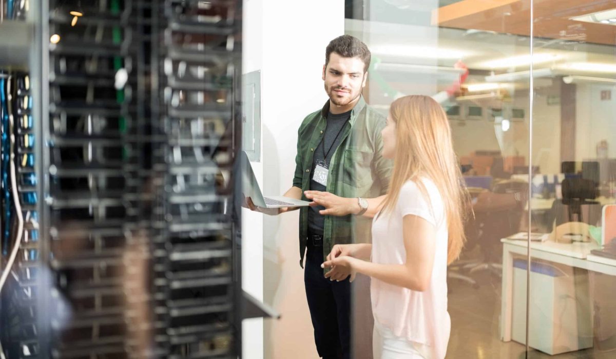 Two people stand near a glass wall in an office, discussing something about the Corvid HPC partnership while one holds a laptop next to server racks.