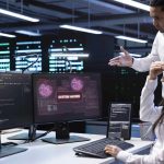 Two IT professionals look at computer screens in a server room; one monitor displays a "SYSTEM HACKED" alert in red, highlighting a cybersecurity breach that may require digital forensics expertise.
