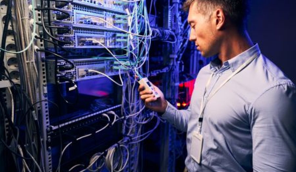 A man in a blue shirt checks a handheld device while standing next to a server rack filled with cables and networking equipment in an HPC data center, possibly monitoring systems powered by NVIDIA RTX PRO 6000 graphics.