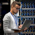 A man in a gray cardigan uses a laptop while standing in front of a server rack filled with numerous cables, showcasing the efficiency of Ace Computers CCS-2 2019B products in a data center.