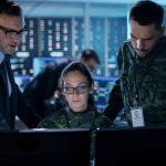 Three individuals working together at computer screens in a government control room equipped with Artificial Intelligence and Cybersecurity technology, against a backdrop of monitors and data servers.