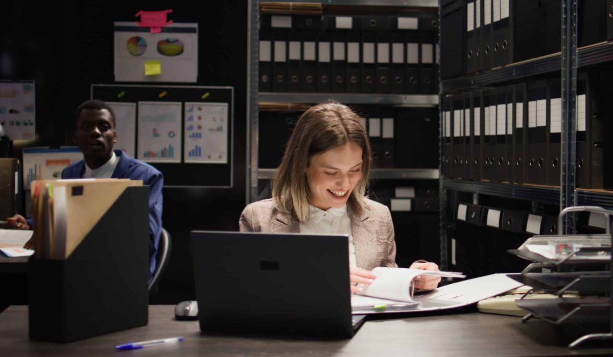 Two people work at desks in an office with shelves full of binders; one woman smiles as she reads a document, while the other uses a computer with a Tableau Write Blocker attached, ensuring data integrity in the background.