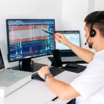 Man wearing headset points at a computer monitor displaying day trading financial charts and graphs, with additional laptops and monitors on desk.