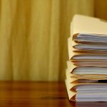 A stack of folders filled with documents is placed on a wooden surface with a yellow curtain in the background.