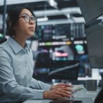 Asian,Woman,In,Headset,Analyzes,Financial,Charts,On,Multiple,Monitors
