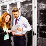 Two professionals stand in a server room, reviewing data on a tablet together, showcasing the advanced technology behind the Corvid HPC partnership, with server racks and networking equipment visible in the background.