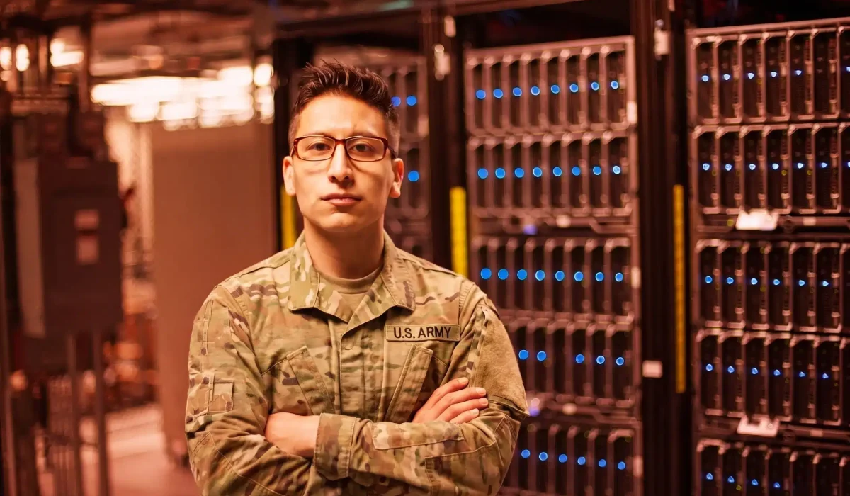 A person in a U.S. Army uniform stands with arms crossed in front of server racks in a federal or military data center.