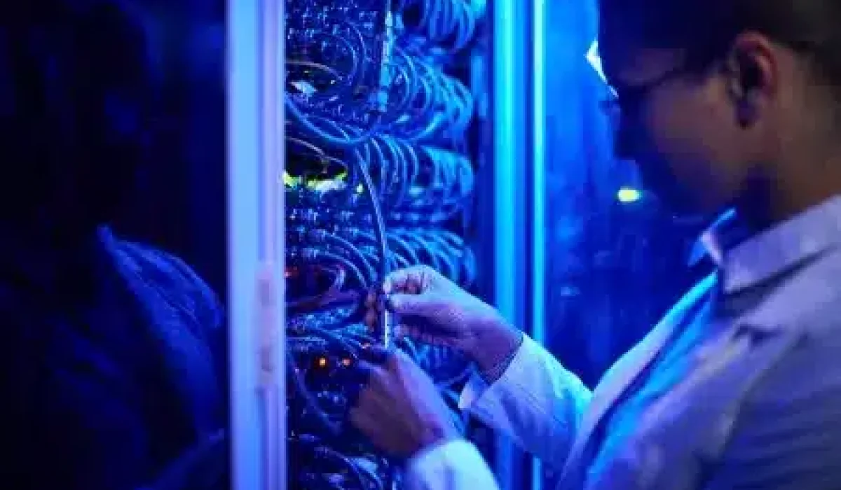 Person in a lab coat adjusts cables inside a server rack in a dimly lit, blue-toned server room featuring traditional networking hardware.